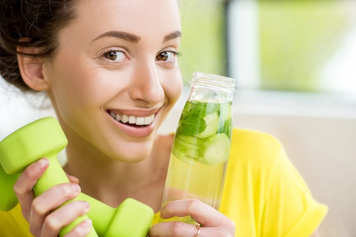 Kobieta pije wodę z miętą, ogórkiem i limonką podczas treningu z hantlami. Koncepcja diety detoksykacyjnej