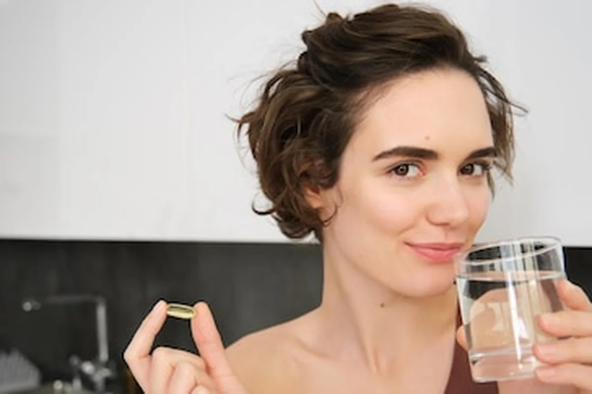 Młoda kobieta przyjmująca suplementy diety i dbająca o zdrowy styl życia z witaminą C, D, Omega, przy szklance wody