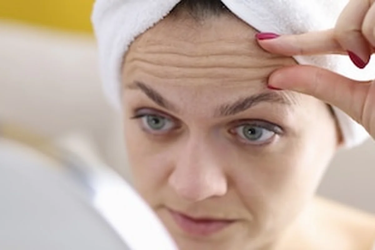 Kobieta bada zmarszczki na czole w lustrze, jak zapobiegać głębokim zmarszczkom mimicznym i grawitacyjnym